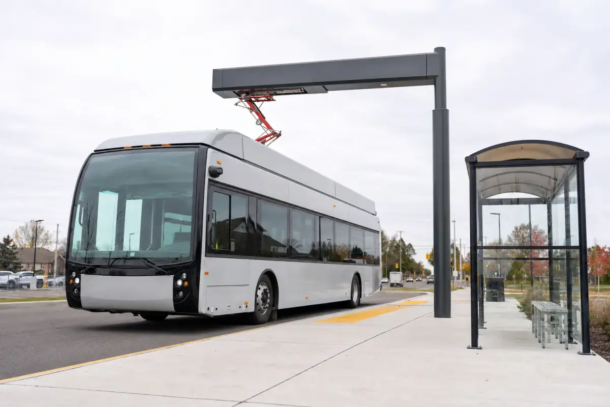 AI illustration: a modern battery-electric transit bus with no markings parked at a clean, empty bus stop shelter, with its charging connector plugged into an overhead pantograph charger above the roof, and a quiet suburban street blurred behind