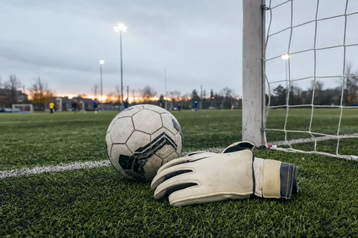 AI illustration: close-up of a pair of soccer goalkeeper gloves and a size-five soccer ball resting on the wet grass of an outdoor turf field beside the white painted line of the goal area, with a softly blurred goal net and floodlight poles behind