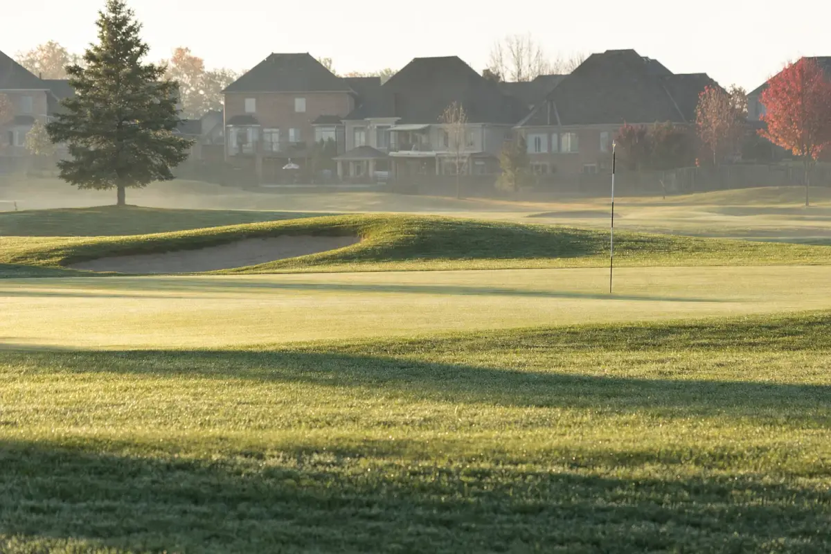 AI illustration: a golf course fairway seen from a low angle on a quiet early morning, with a neatly mown green, a single flagstick with no flag markings and a sand bunker in the middle distance, backed by the rear fences and rooftops of a few suburban houses softly blurred