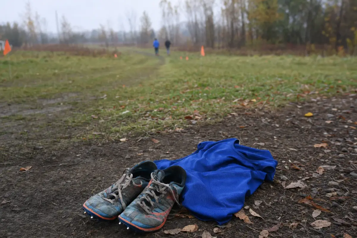 AI illustration: close-up of a pair of worn running spikes and a plain pinned-free cotton race bib-less singlet resting on a dirt trail edge at a cross country course, with a worn grassy trail and a few orange course flags blurred behind