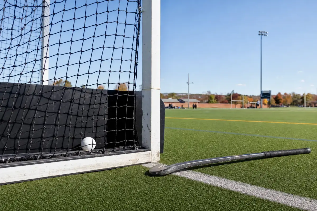AI illustration: close-up of a field hockey goal on an outdoor turf field, with a white ball resting just inside the back of the net and a curved stick lying on the green turf in front of the goal line