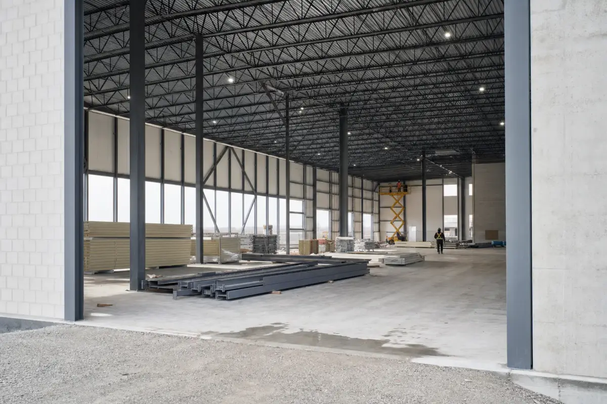 AI illustration: interior of a large, bright industrial manufacturing hall under construction, with a bare poured concrete floor, tall steel columns and roof trusses, stacks of plain steel beams and pallets of insulation panels, and a yellow scissor lift parked near the back