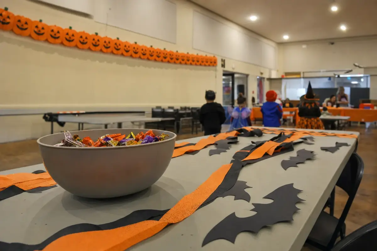 AI illustration: inside a bright community hall set up for a family Halloween event, with a long folding table of plain paper bats and orange and black streamers, a big plain bowl of wrapped candies and a few small costumed children's silhouettes blurred in the background