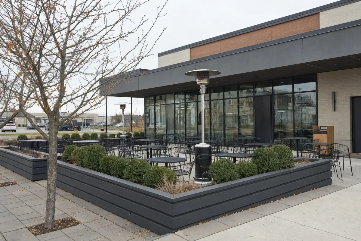 AI illustration: a restaurant patio on a quiet suburban commercial strip just before opening, with empty black metal tables and chairs, a low planter fence and a couple of unlit patio heaters, with a plain modern single-storey building with large windows blurred behind
