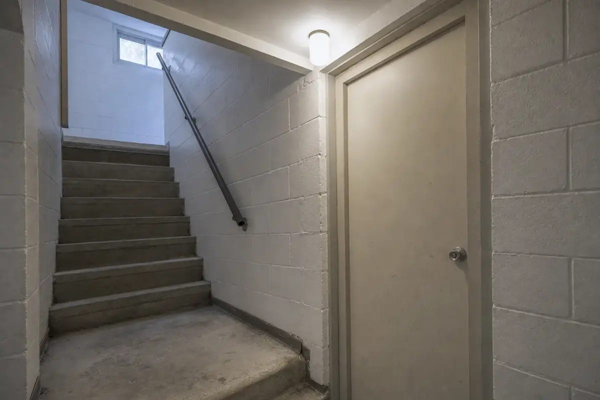 AI illustration: close-up of a plain interior door at the bottom of a basement stairwell in an older low-rise residential building, with a bare concrete step, a metal handrail, a clean painted cinder-block wall and a small plain light fixture overhead