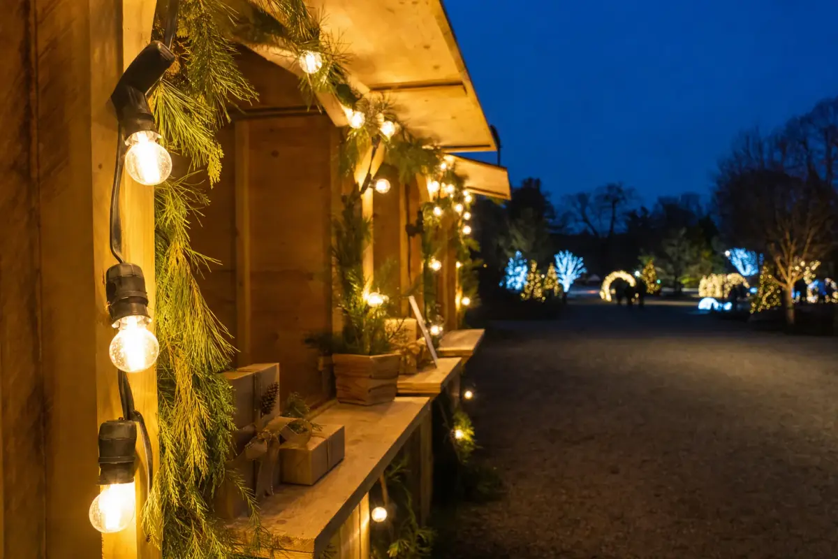 AI illustration: close-up of a strand of warm white string lights and a plain wooden market stall with a few unbranded wrapped gift boxes and evergreen boughs, set along a dark garden path, with softly glowing light displays blurred in the distance
