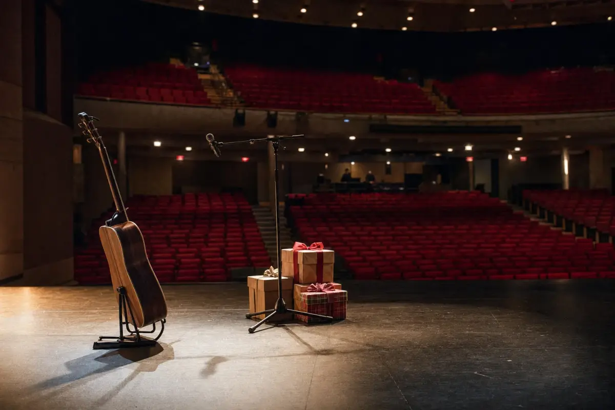 AI illustration: interior of a empty concert theatre seen from the side of the stage, with a lone acoustic guitar on a stand and a microphone beside a small plain stack of wrapped gift boxes, rows of red velvet seats and a balcony blurred beyond