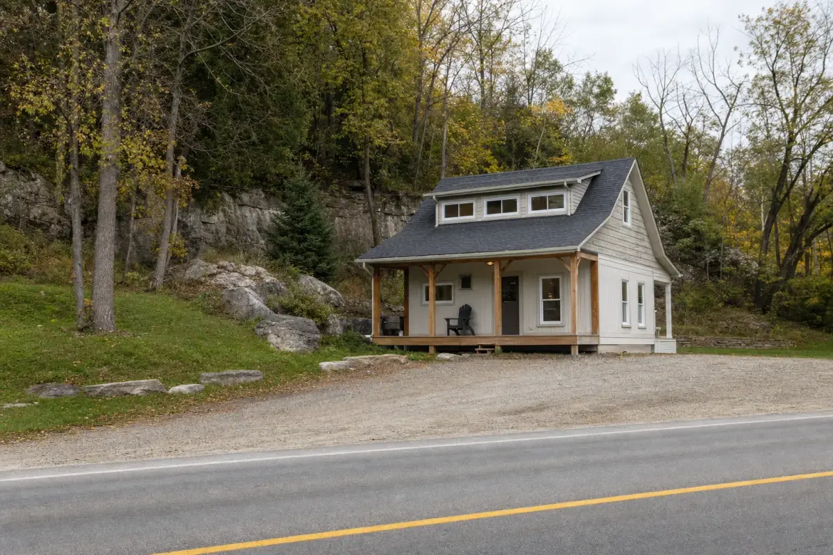 AI illustration: a rustic, clean country house with a small wooden porch and a plain gravel driveway set back from a rural road on a wooded escarpment slope, with a few rocky outcrops and mature trees behind and an empty patio chair by the door