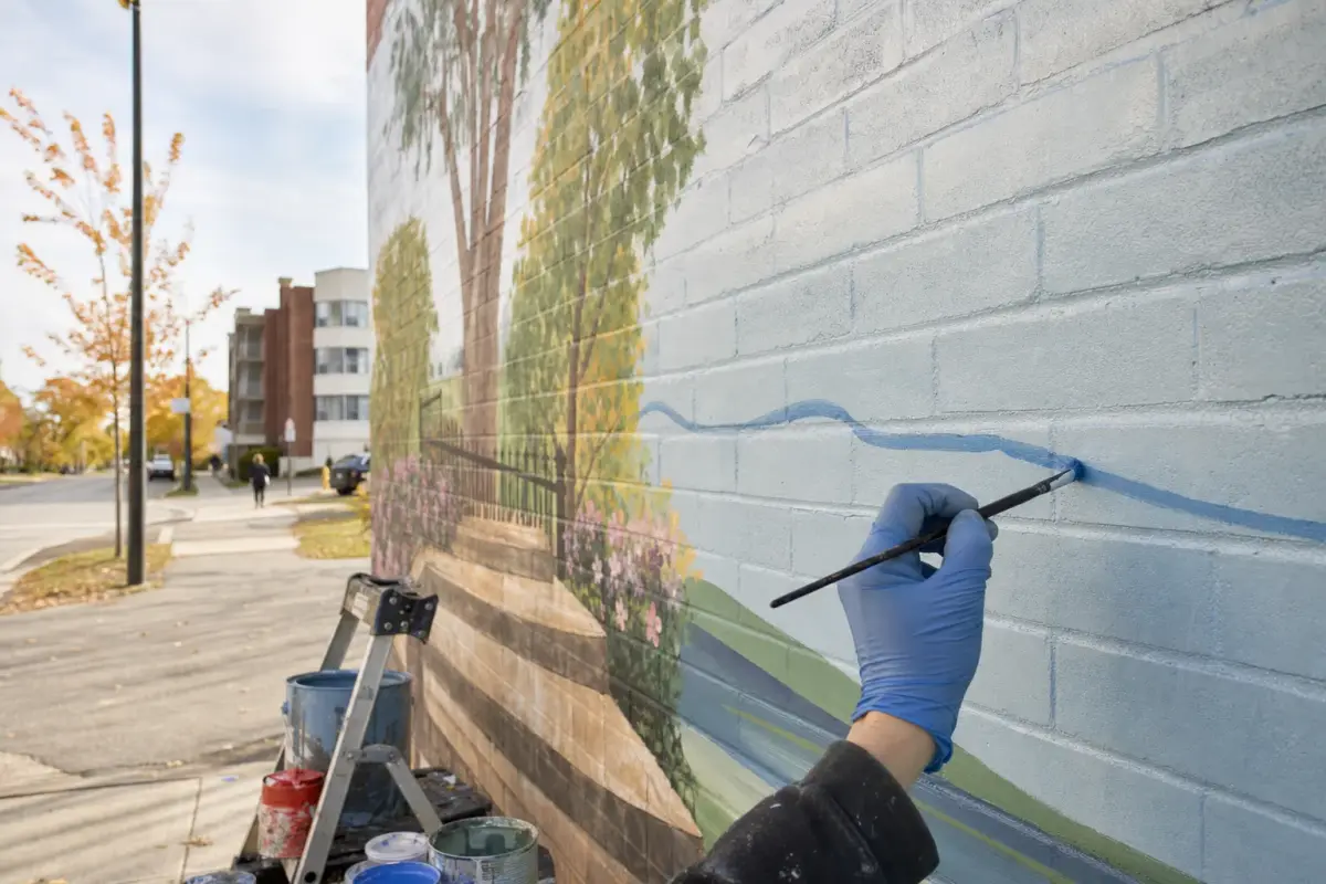 AI illustration: close-up, slightly low angle of a painter's gloved hand holding a fine brush against a large brick wall, adding a soft blue wave line to a half-finished colourful landscape mural, with open paint tins and a stepladder at the base