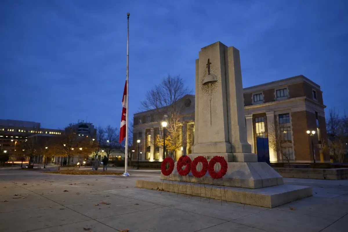 AI illustration: a stone war memorial cenotaph at the edge of a quiet civic plaza at dusk, with a few plain red wreaths on wire stands resting at its base and a flagpole with a limp flag to one side, and a softly blurred municipal building with a few lit windows behind