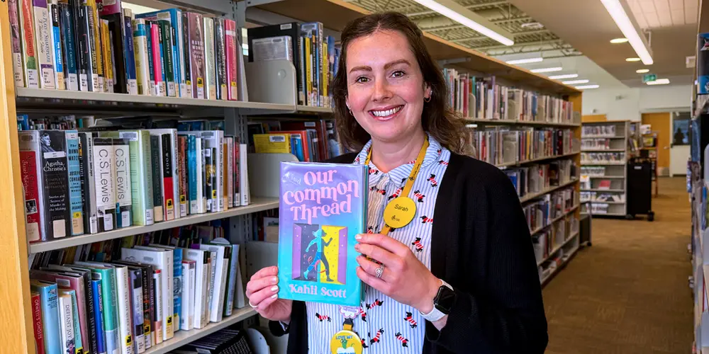 Librarian Sarah stands in front of book stacks holding the book Our Common Thread.