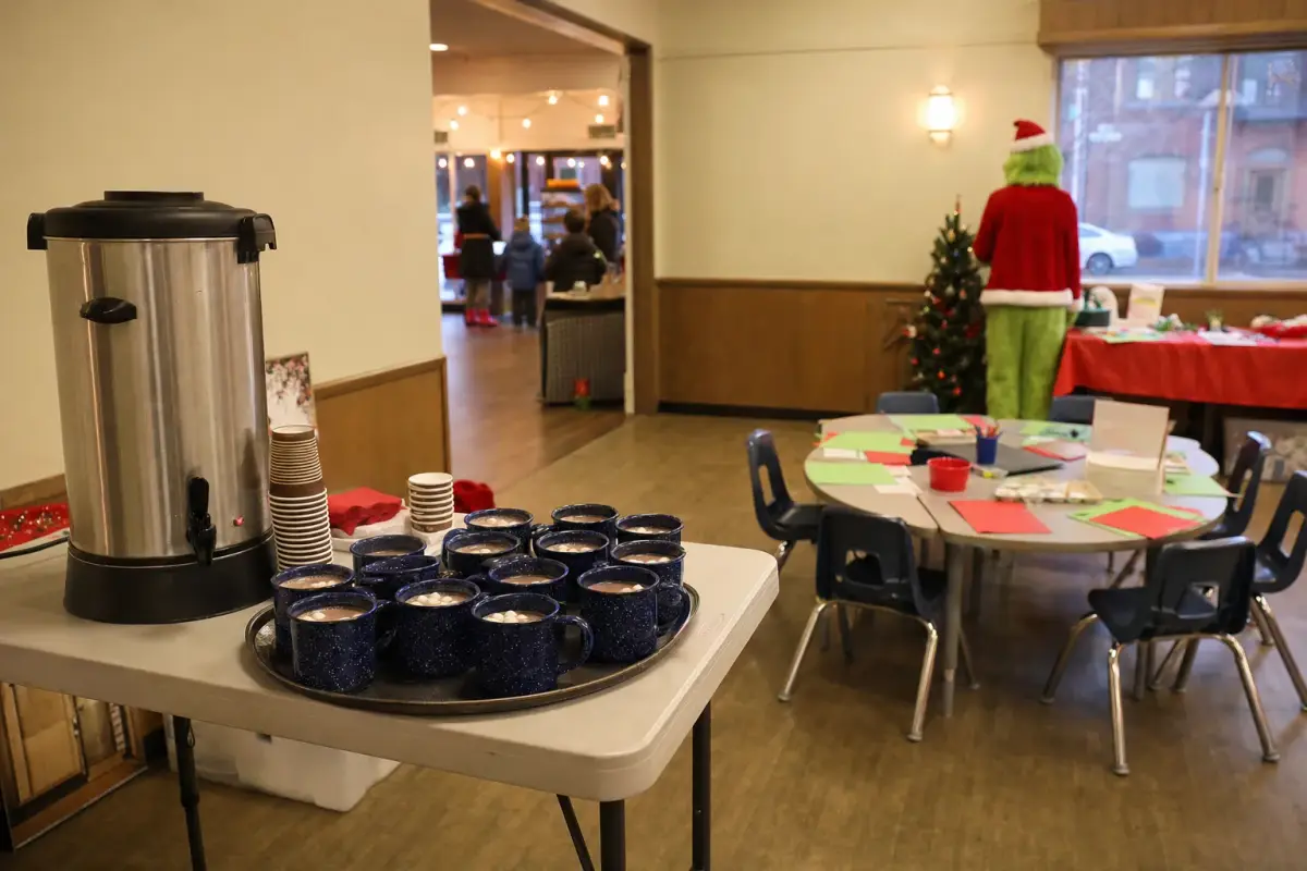 AI illustration: interior of a community hall set up as a warm-up pop-up, with a folding table holding a large steel urn, a stack of paper cups and a tray of hot cocoa mugs, a table of plain paper crafts with red and green construction paper and child-sized chairs beside it, and a softly blurred window showing a darkening street outside