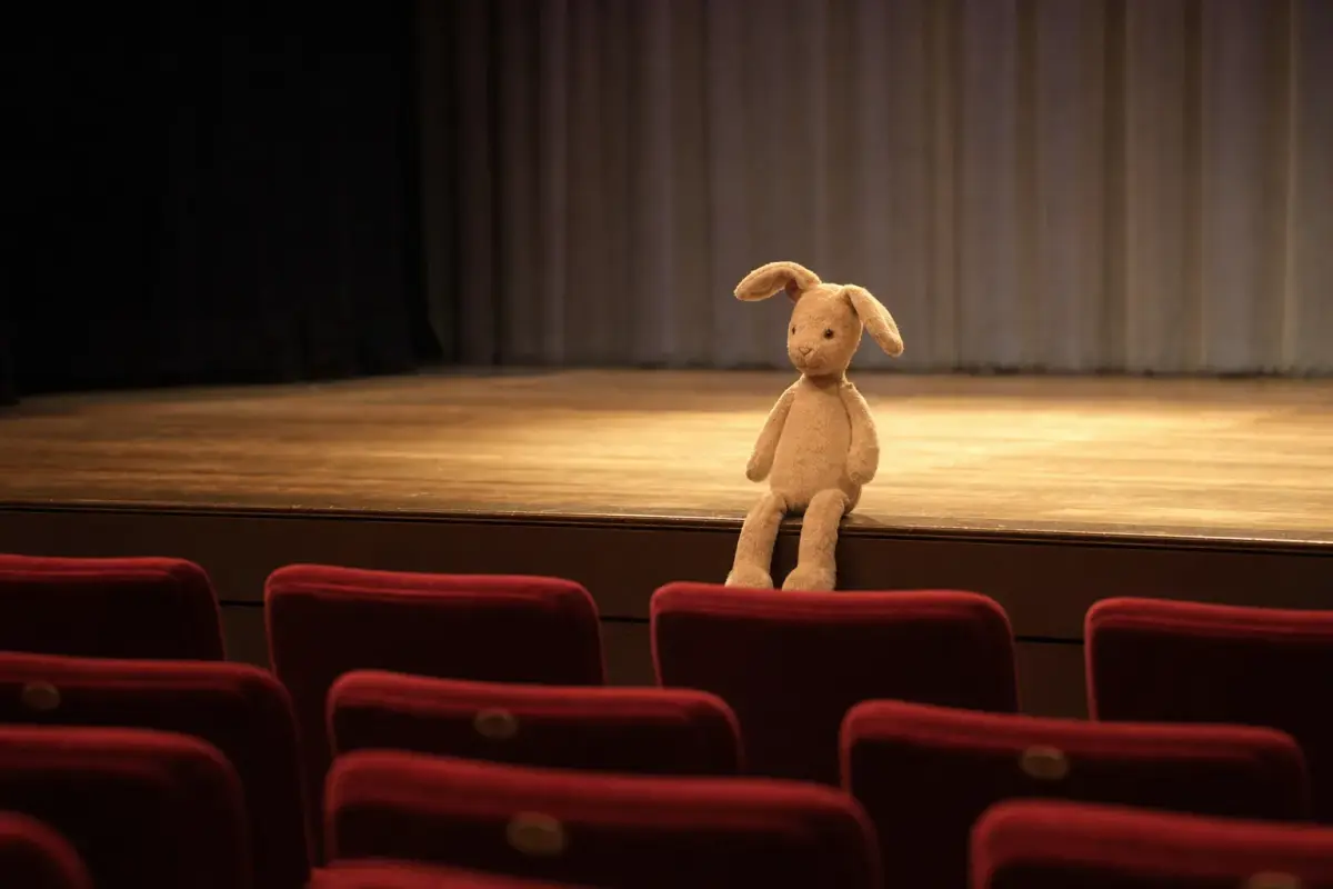AI illustration: interior of a small theatre auditorium before a children's matinee, with rows of empty red upholstered seats, a plain stuffed toy rabbit sitting on the edge of the low stage, and a soft pool of stage light on bare wooden boards, with a plain curtain blurred behind